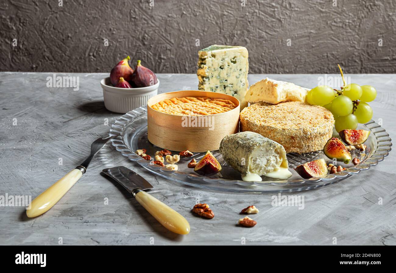 Auswahl an französischem Käse - Camembert, roquefort, Brie, Ziegenkäse und Epoisse mit Trauben, Feigen und Nüssen auf grauem Hintergrund Stockfoto