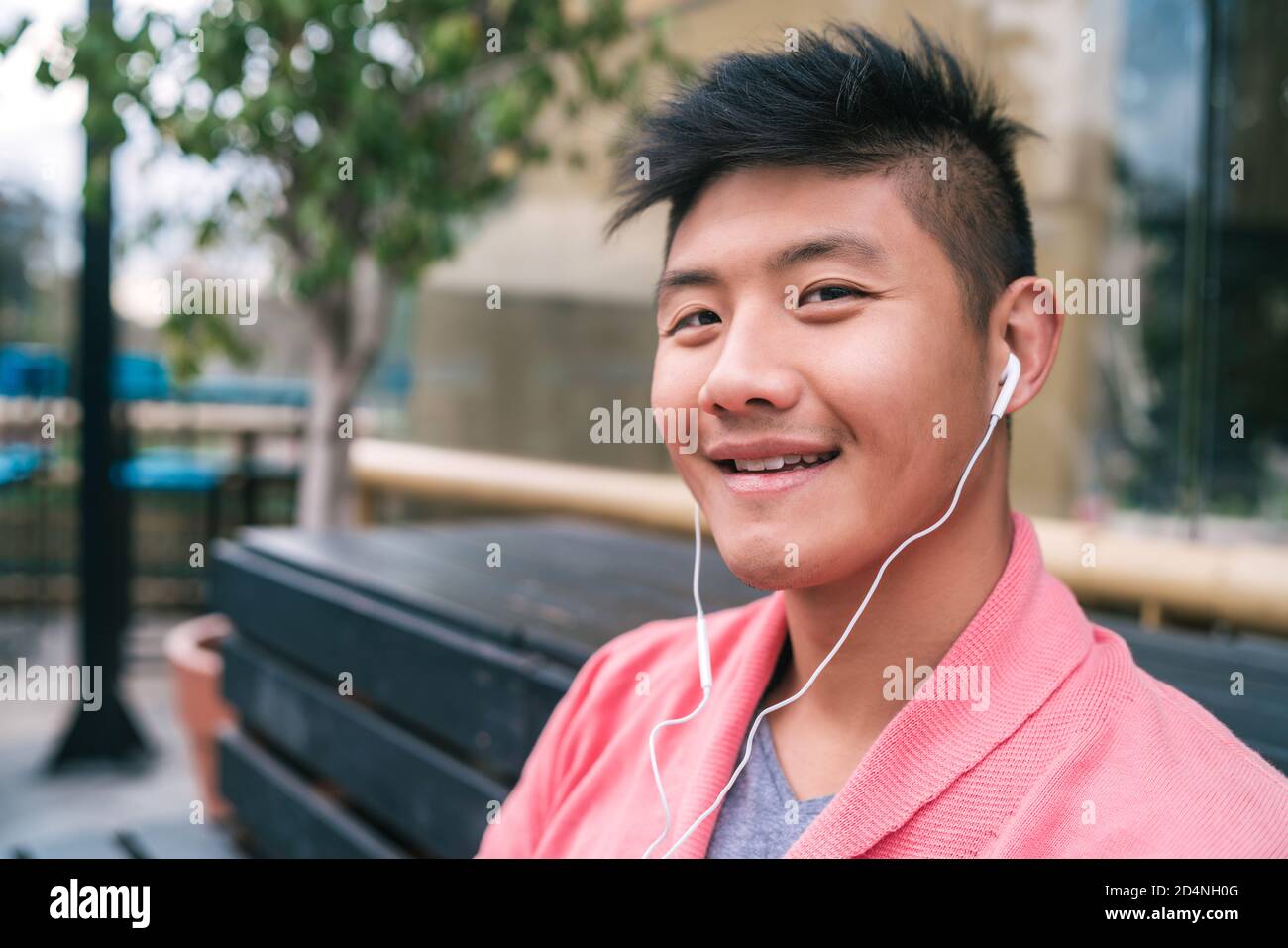 Porträt eines jungen asiatischen Mannes, der mit Kopfhörern Musik hört, während er auf einer Bank in einem Park sitzt. Stockfoto