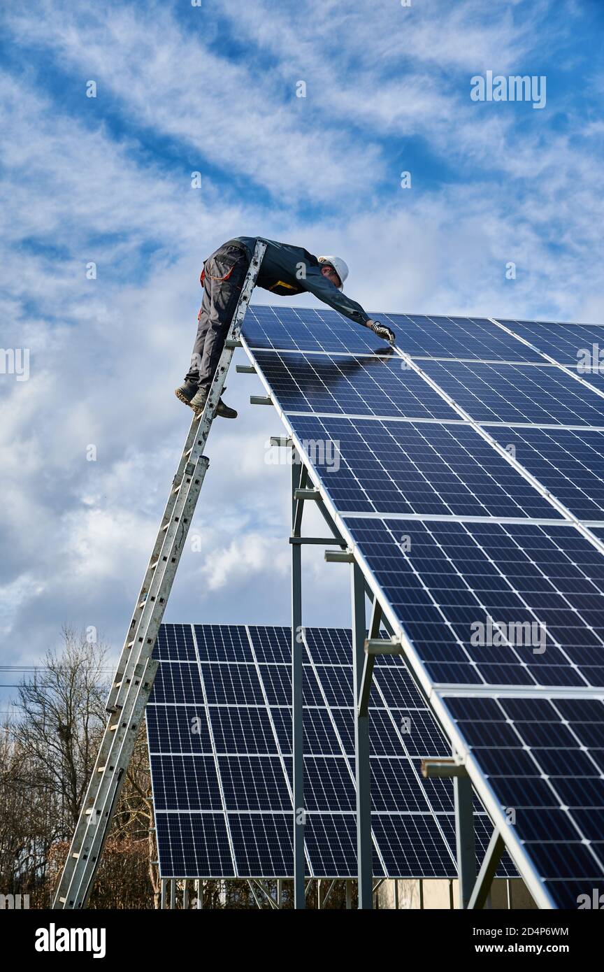 Vertikale Aufnahme von Mann Arbeiter Elektriker Reparatur Photovoltaik-Solarmodul, stehend auf Leiter unter schönen bewölkten Himmel, Installation Solar-Photovoltaik-Panel. Konzept der alternativen Energie Stockfoto