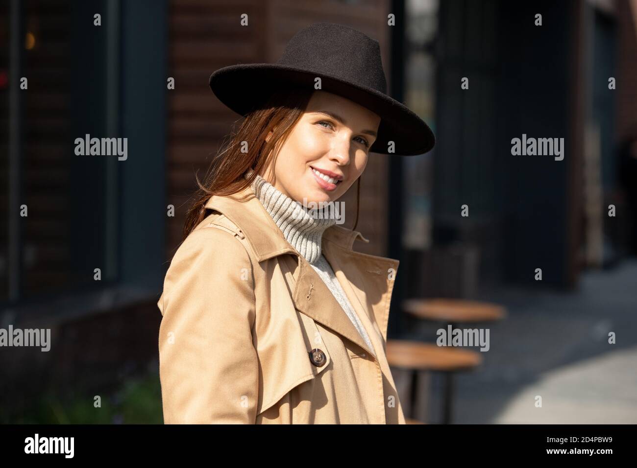 Glückliche junge wunderschöne Frau in schwarzem Hut, beigem Trenchcoat und grauem Pullover Stockfoto