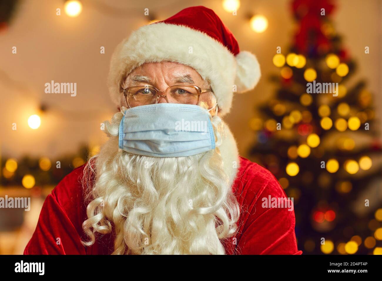 Santa mit einem Bart in einer Schutzmaske auf seinem Gesicht zu Weihnachten gekleidet schaut auf die Kamera. Bleiben Sie zu Hause. Stockfoto