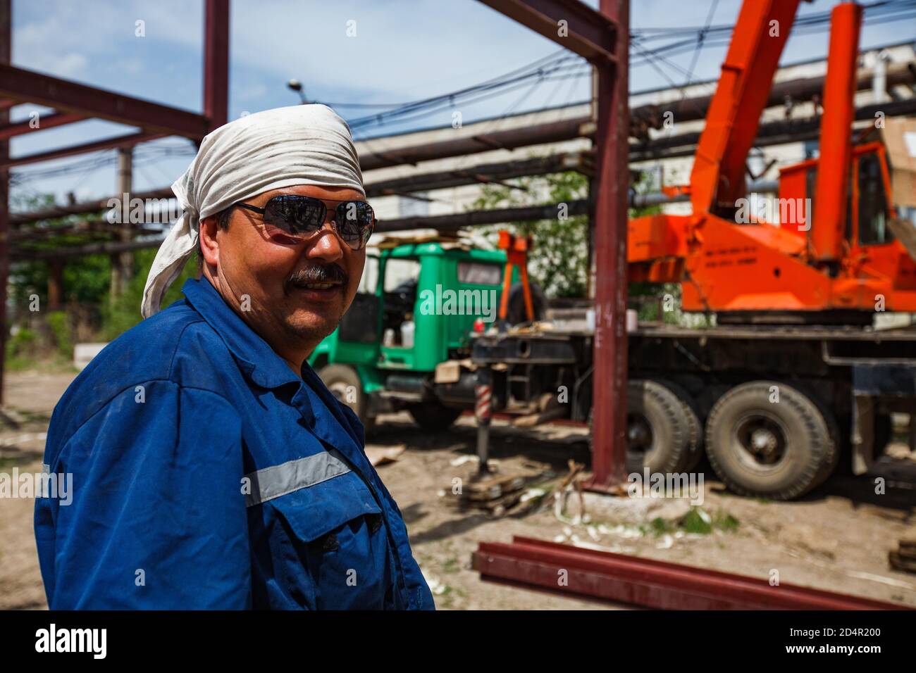 Shymkent/Kasachstan - April 27 2012: Porträt eines usbekischen Arbeiters in Sonnenbrille und weißem Stoffkopfdeckel. Orangefarbener und grüner Mobilkran auf dem Hintergrund. Stockfoto