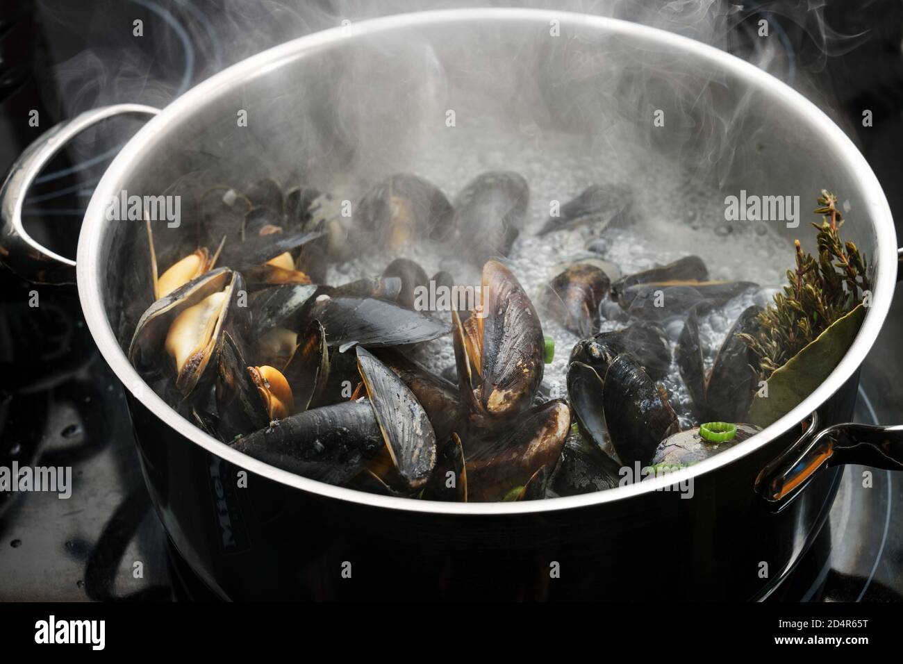 Dampfende blaue Muscheln in einem Stahltopf mit kochender Brühe, Zwiebeln und Kräutern auf einem schwarzen Herd, Kochen ein köstliches Meeresfrüchte-Rezept, ausgewählte Fokus, schmal Stockfoto