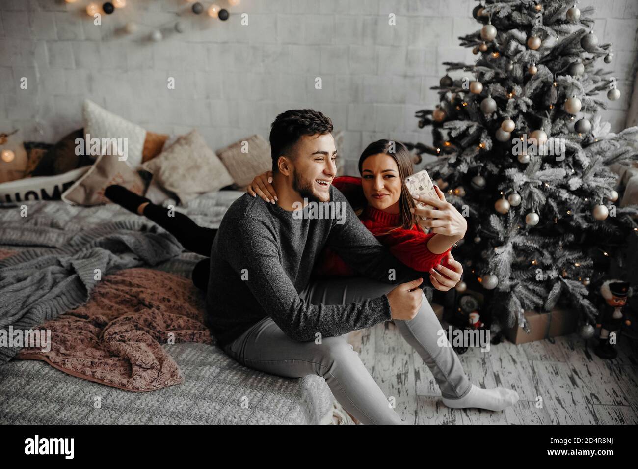 Glückliches junges Paar, das zu Hause Selfie macht, Spaß hat, auf einem gemütlichen Bett zu liegen und neben einem Weihnachtsbaum zu lachen Stockfoto