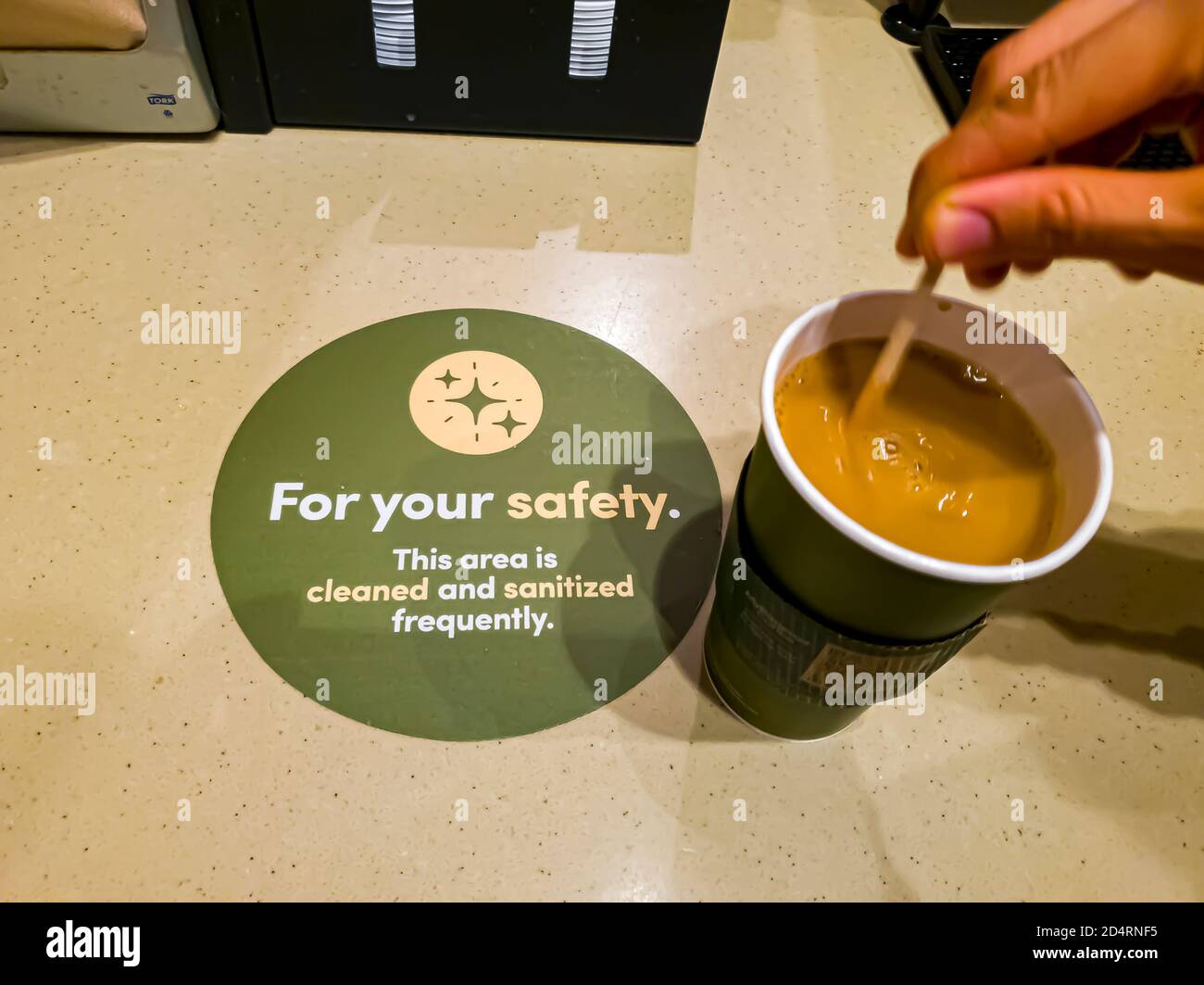 Rockville, MD, USA 10/09/2020: Eine Frau mischt und rührt Kaffee auf der Bank in einem Café. Es gibt einen Hinweis mit der Aufschrift "zu Ihrer Sicherheit, dies ar Stockfoto