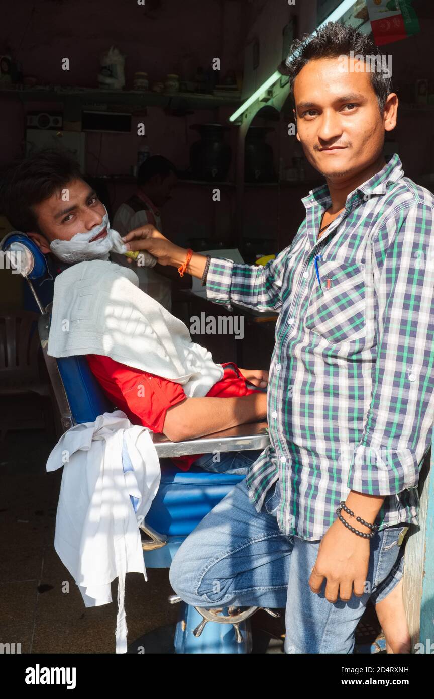 Ein Barbier in Mumbai, Indien, und ein Kunde in dem kleinen, offenen Friseurladen, sein Gesicht in Rasiercreme eingewaschen, posieren für den Fotografen Stockfoto