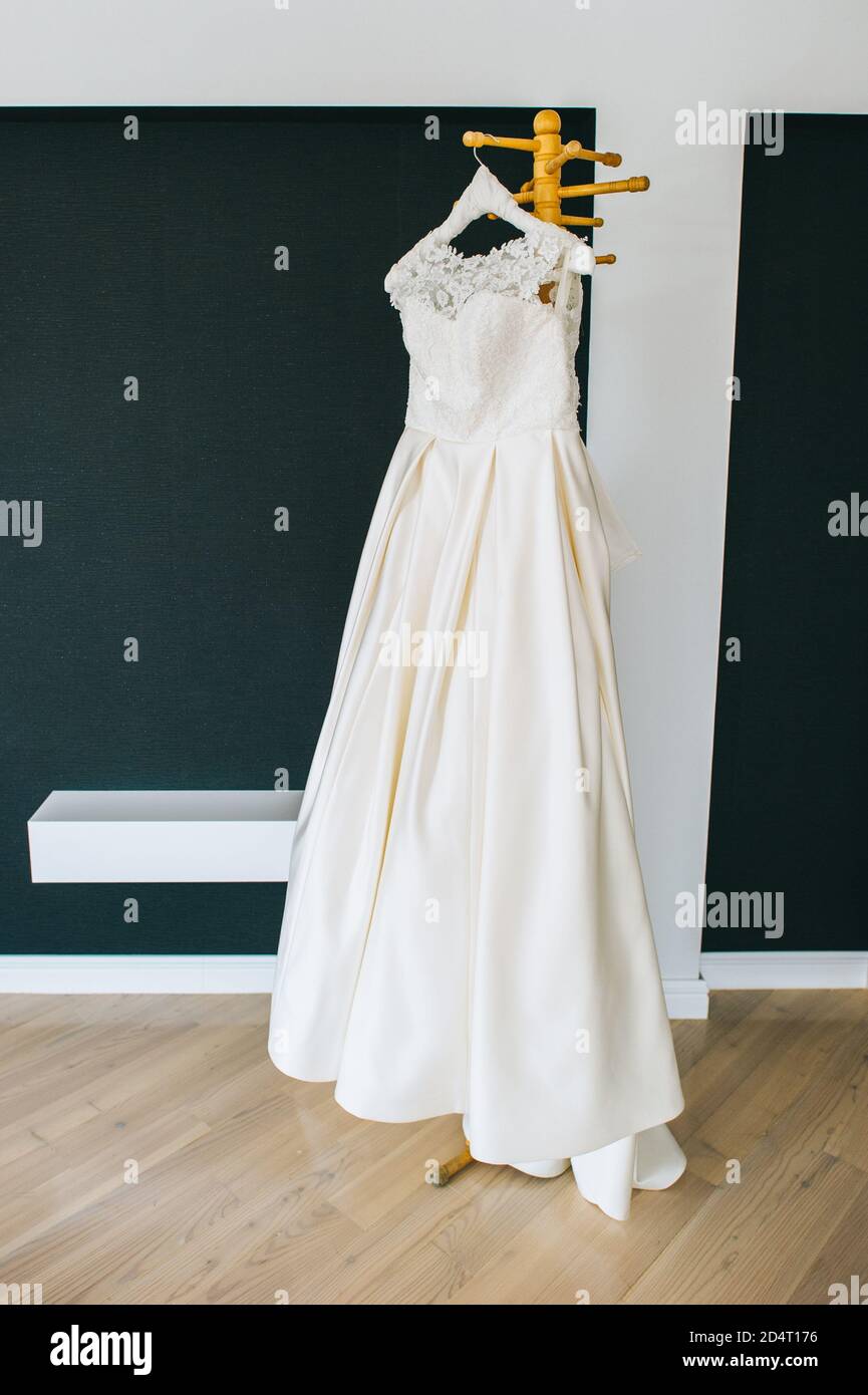 Schönes weißes minimalistisches Hochzeitskleid, das auf einem Holzhänger in der Mitte des Raumes hängt. Stockfoto