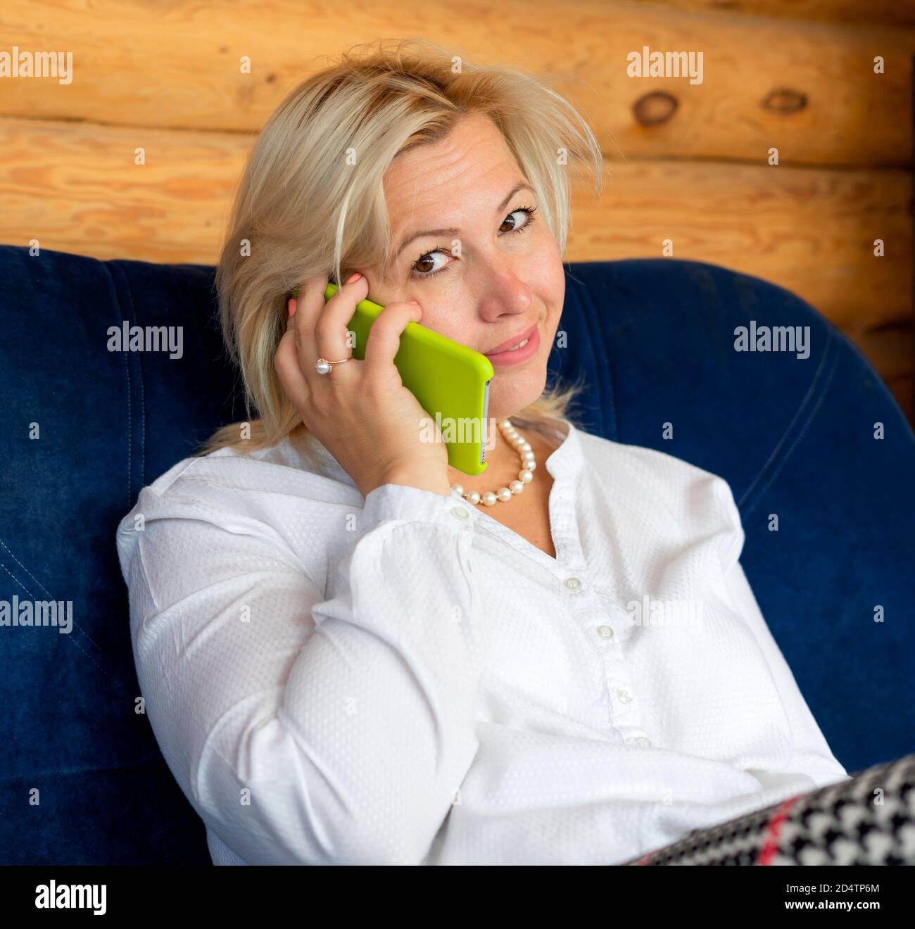 Eine Blondine mittleren Alters in einem hölzernen Landhaus spricht am Telefon mit einem Lächeln. Arbeiten Sie von zu Hause aus. Remote-Arbeit. Stockfoto