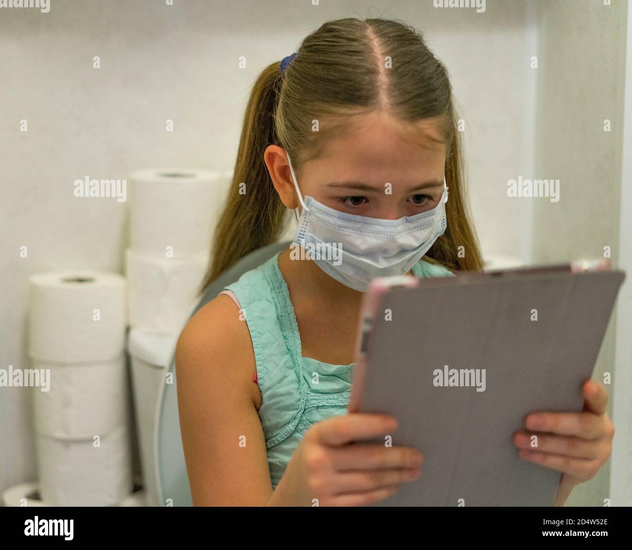 Ein trauriges Mädchen in einer medizinischen Maske mit einer Tablette in den Händen sitzt in der Toilette. Stockfoto