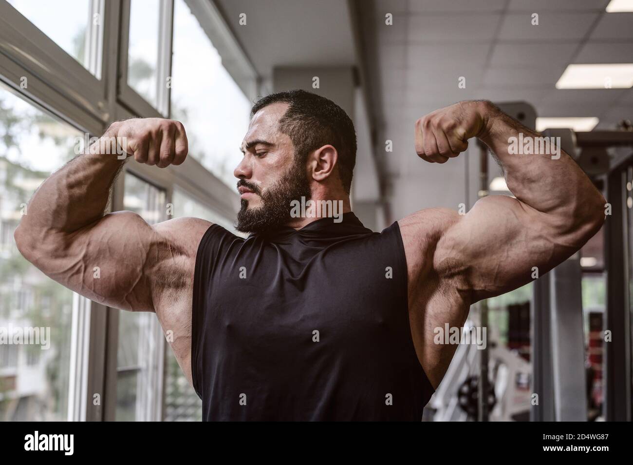Starker junger Athlet Mann mit Bart trägt schwarzen Tank Top Zeigt große doppelte Bizeps Muskel in Sporthalle mit Fenster Stockfoto