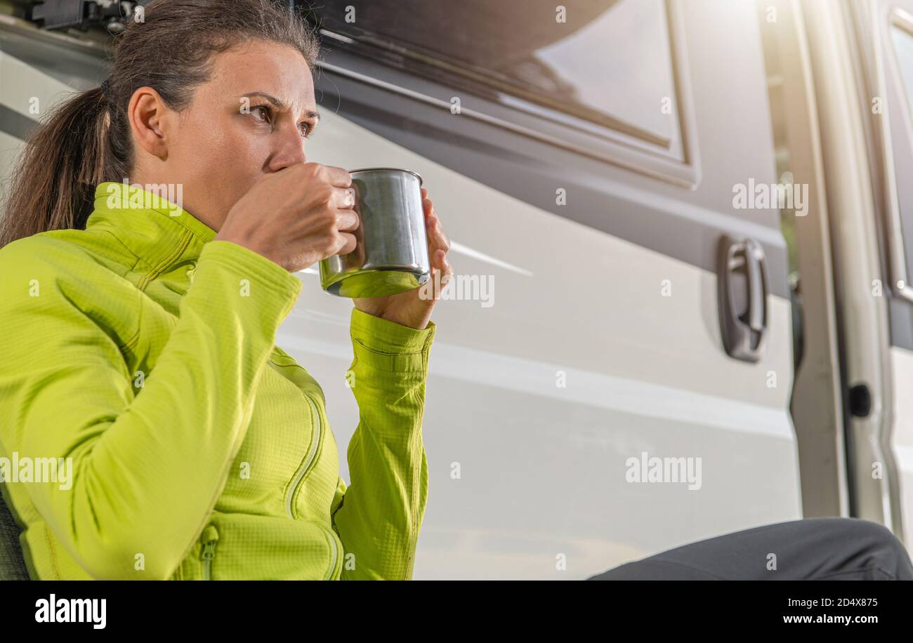 Kaukasische Frau in ihren späten 30er Jahren trinken Tasse frisch Gebrühter Kaffee vor ihrem modernen Camper Van während Camping in Wilderness Stockfoto