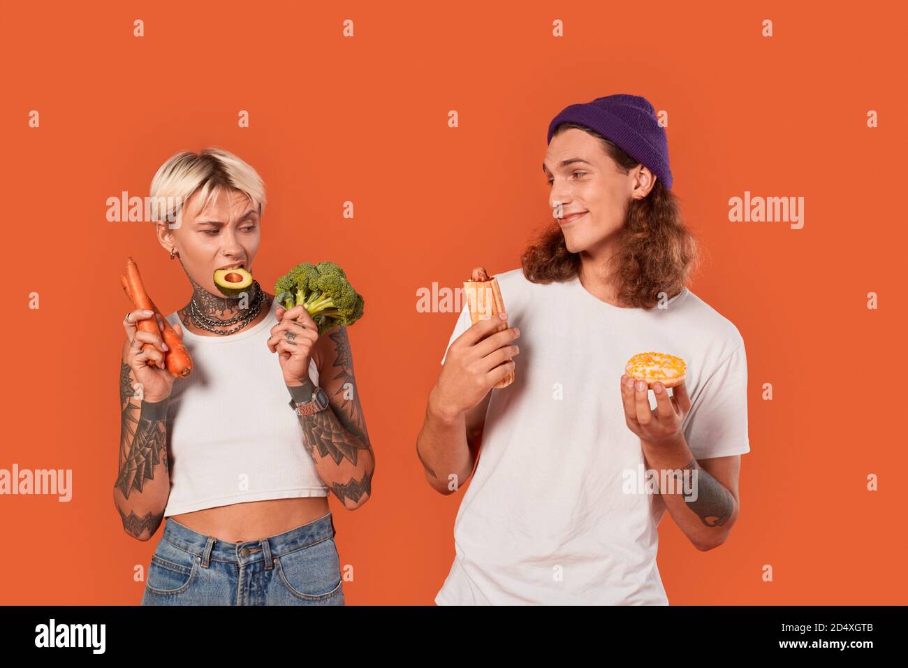 Freistil. Junge tätowierte Paar stehend isoliert auf orange Freundin hält Gemüse gesunden Lebensstil vs Freund hält Hot Dog und Donut Stockfoto