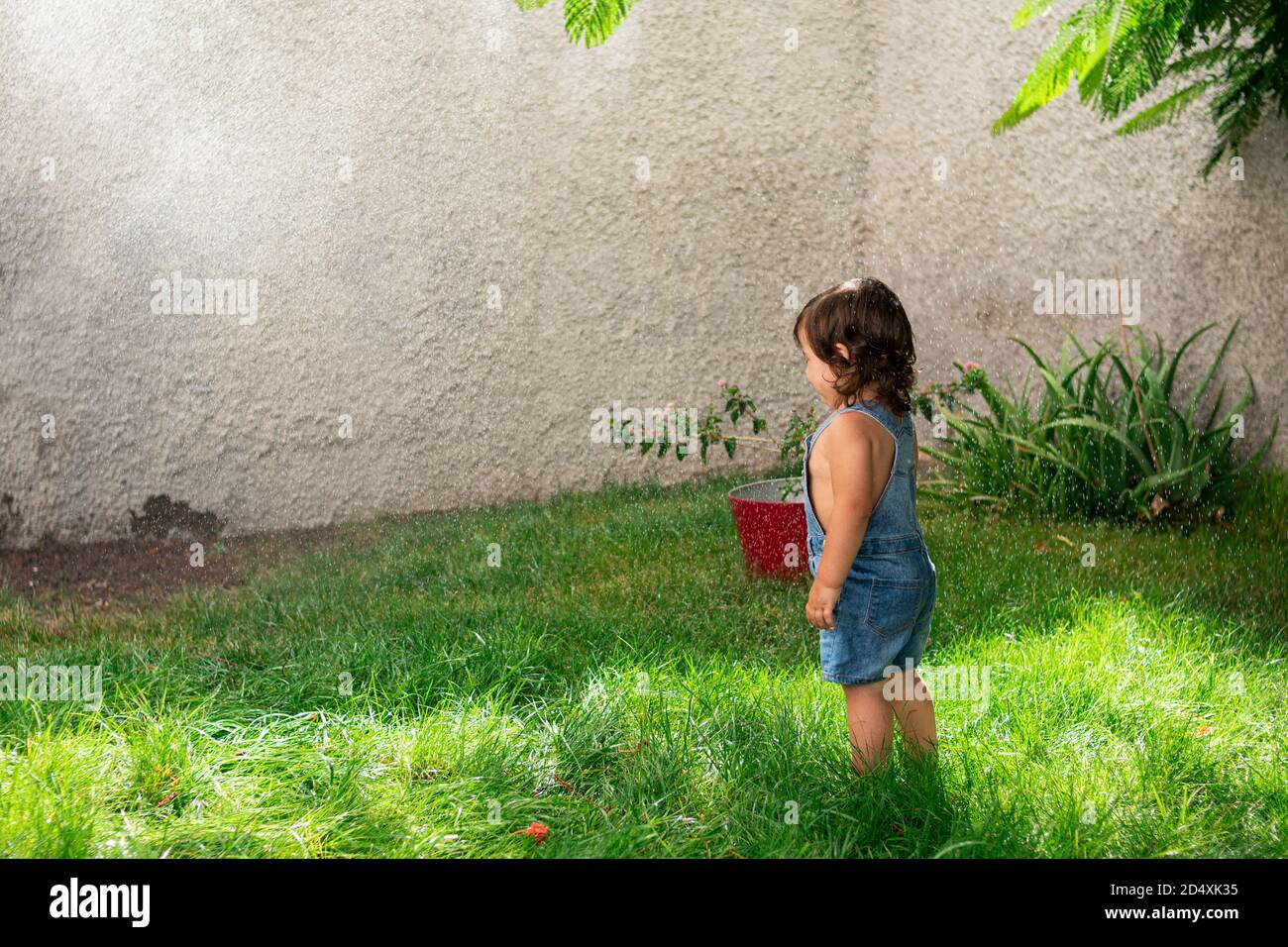 Augenhöhe Aufnahme eines niedlichen Mädchen in insgesamt Denim Shorts unter spritzenden Wasser in einem Garten Stockfoto