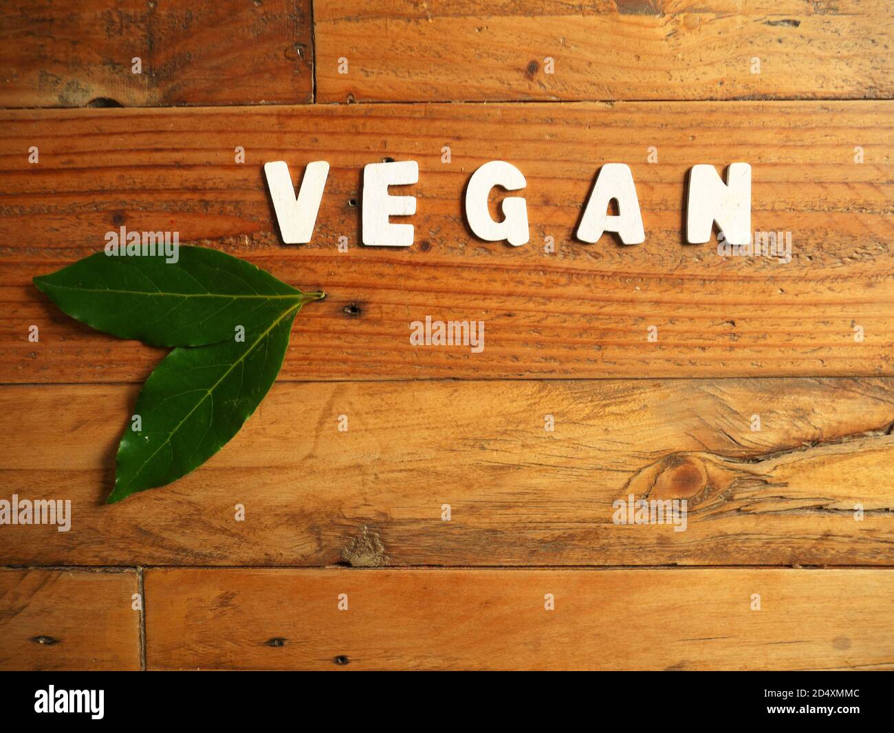 Wort 'VEGAN' in weißen Buchstaben auf einem Holzhintergrund mit Zwei Lorbeerblätter Stockfoto