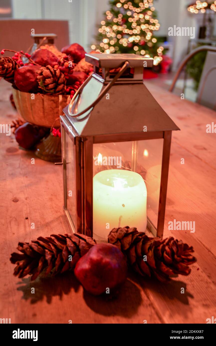 Weihnachtliche Tischdekorationen mit glühender Kerze, Tannenzapfen und Granatapfel als Herzstück, mit einem beleuchteten Weihnachtsbaum im Hintergrund. Stockfoto