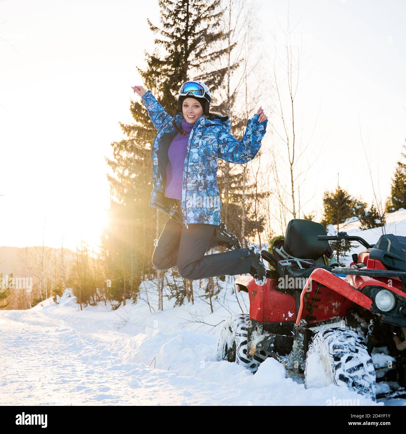 Portrait von schönen blonden Mädchen, trägt blaue Winterjacke Springen neben roten Vierrad ATV im Winter in verschneiten Bergen. Hell untergehende Sonne auf dem Hintergrund. Aktives Lifestyle-Konzept Stockfoto
