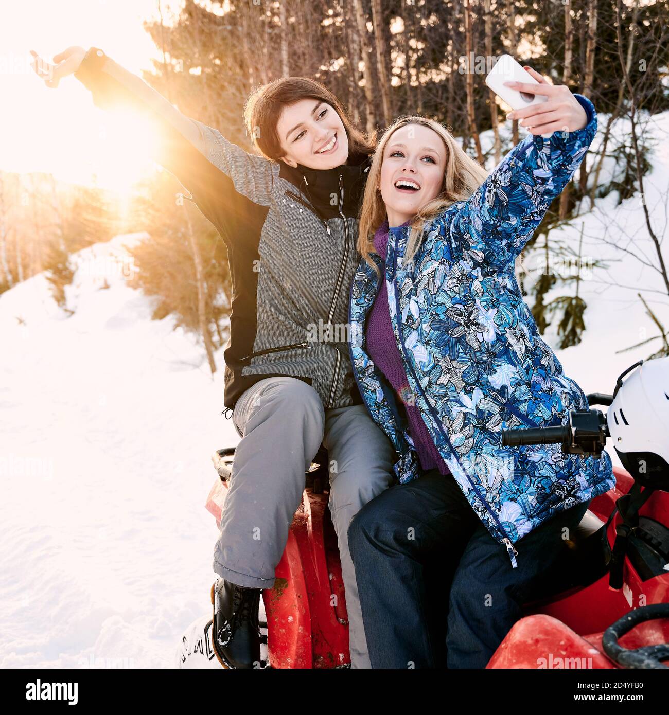 Zwei fröhliche hübsche Mädchen posieren Selfie gegen Sonnenuntergang in verschneiten Bergen sitzen auf roten Quad-Bike. Konzept der Winterunterhaltung Stockfoto