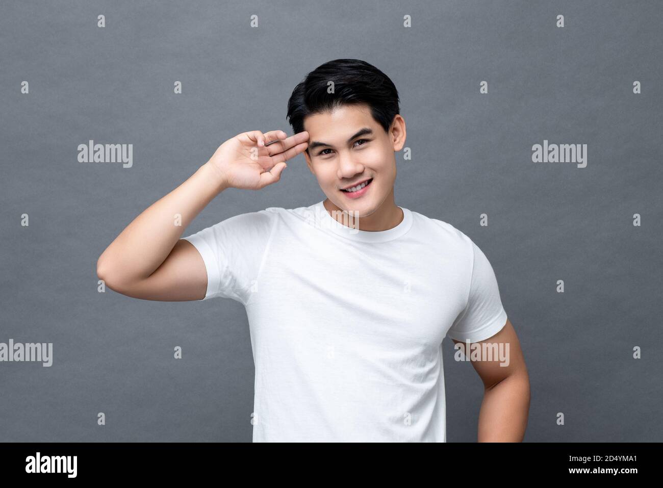 Taille bis Porträt von gut aussehend lächelnd freundlich asiatischen Mann in Lässiges weißes einfarbiges T-Shirt mit Finger-auf-Kopf-Geste isoliert Auf grauem Hintergrund Stockfoto