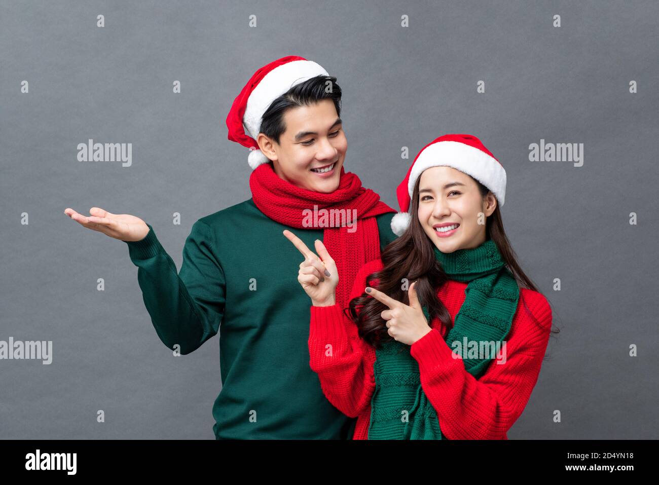 Lächelndes asiatisches Paar in Weihnachtskleidung tun offene Hand und Zeigegegesten für die Produktplatzierung auf grauem Studiohintergrund Stockfoto