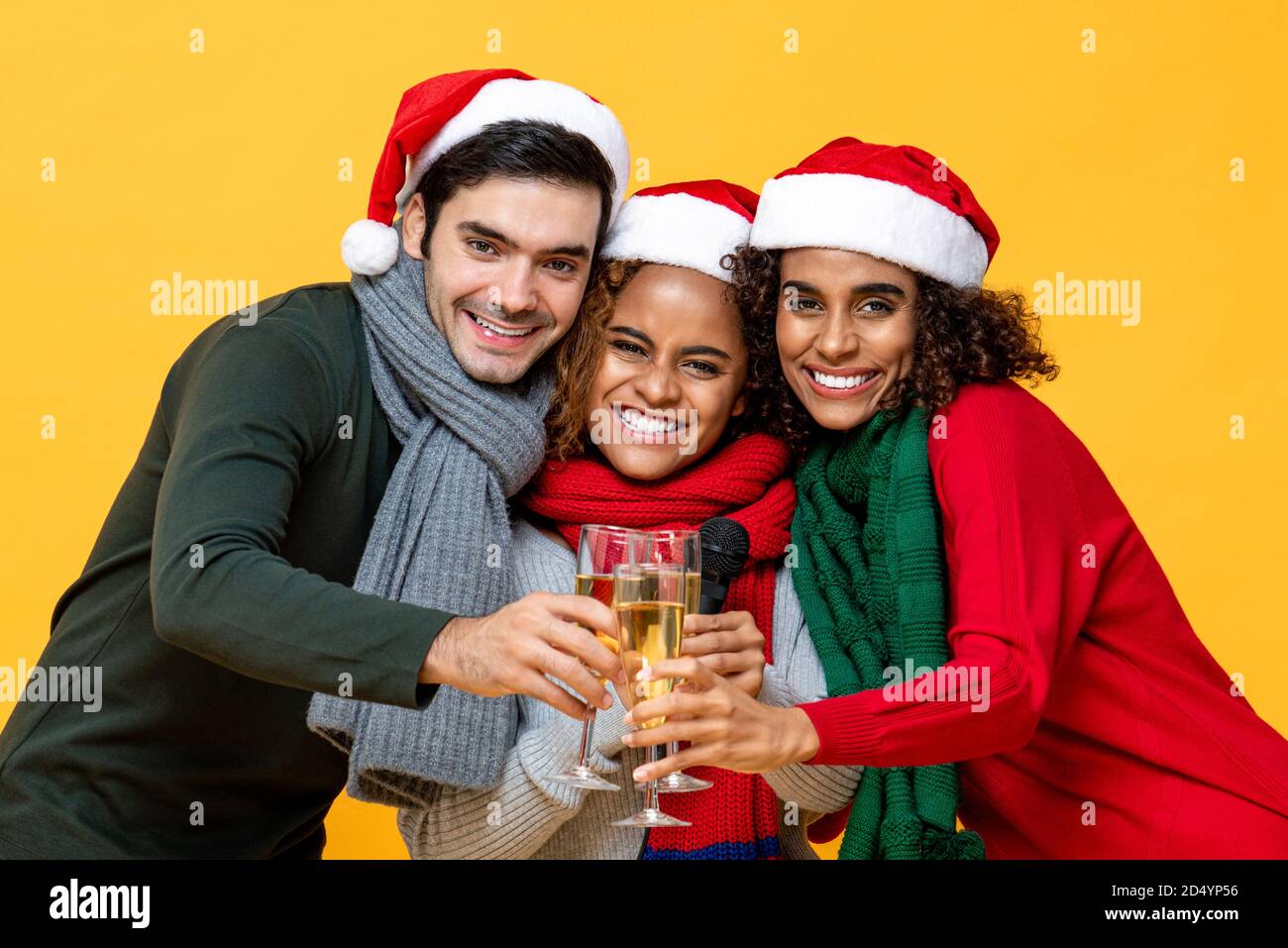 Drei glückliche diverse Freunde in Weihnachtskleidung klirrende Gläser von Champagner feiert im Studio gelben Hintergrund Stockfoto
