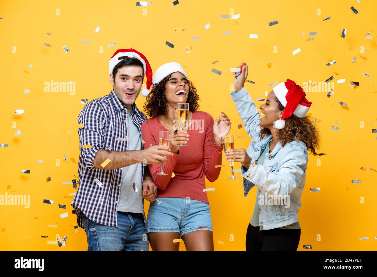 Eine Gruppe von drei glücklichen und vielfältigen Freunden feiert Weihnachten mit Champagner Und tanzen auf gelbem Studiohintergrund mit Konfetti Stockfoto