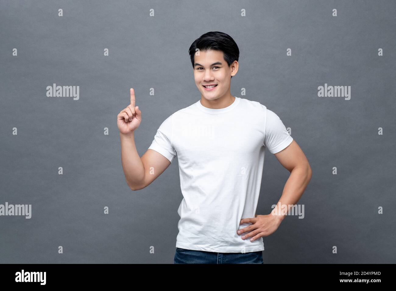 Lächelnd schöner asiatischer Mann poiting Finger bis zu leeren Raum Im Studio grau isoliert Hintergrund Stockfoto