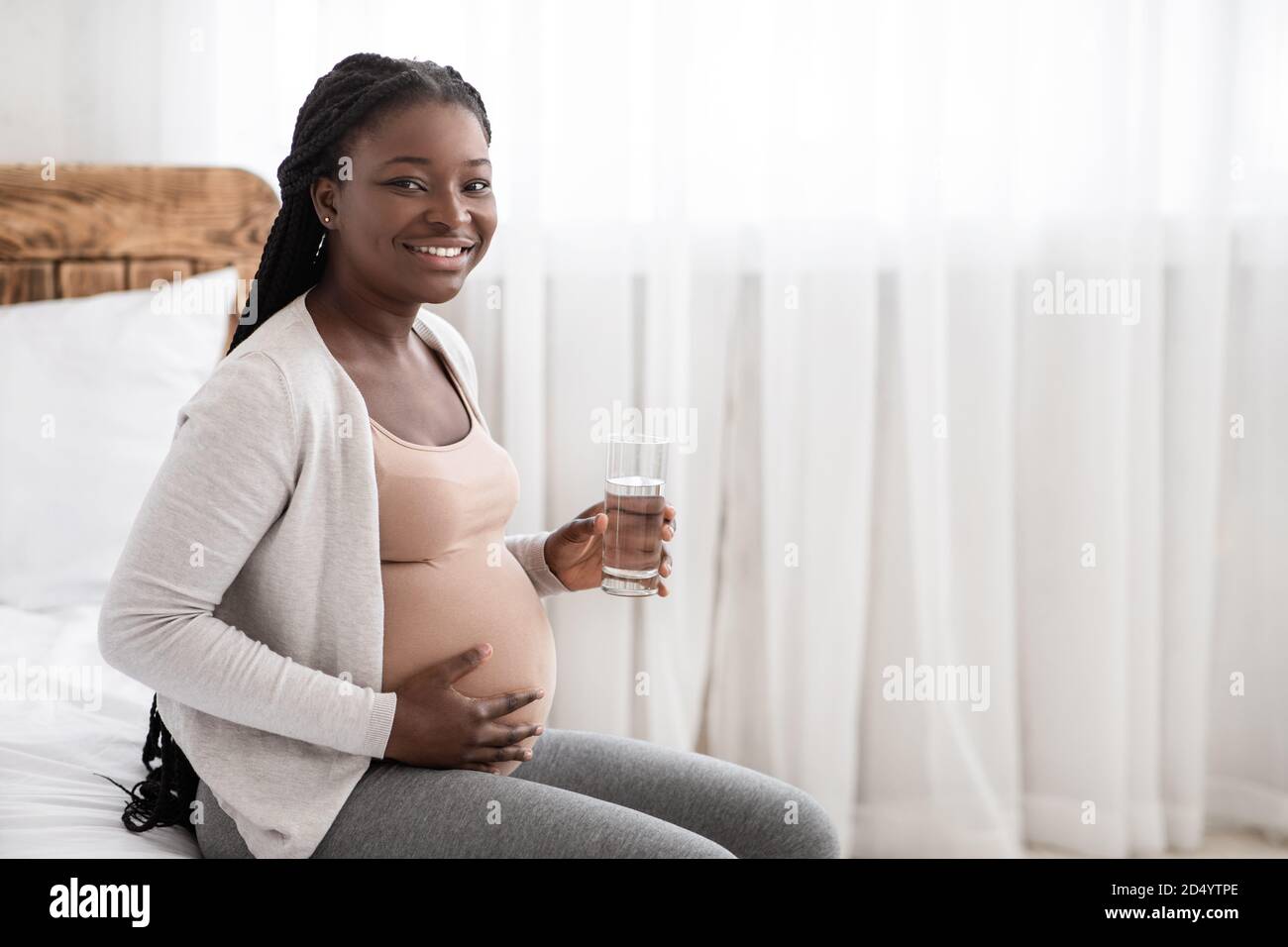 Happy African Schwangere Frau Trinkwasser Aus Glas Beim Entspannen Auf Dem Bett Stockfoto