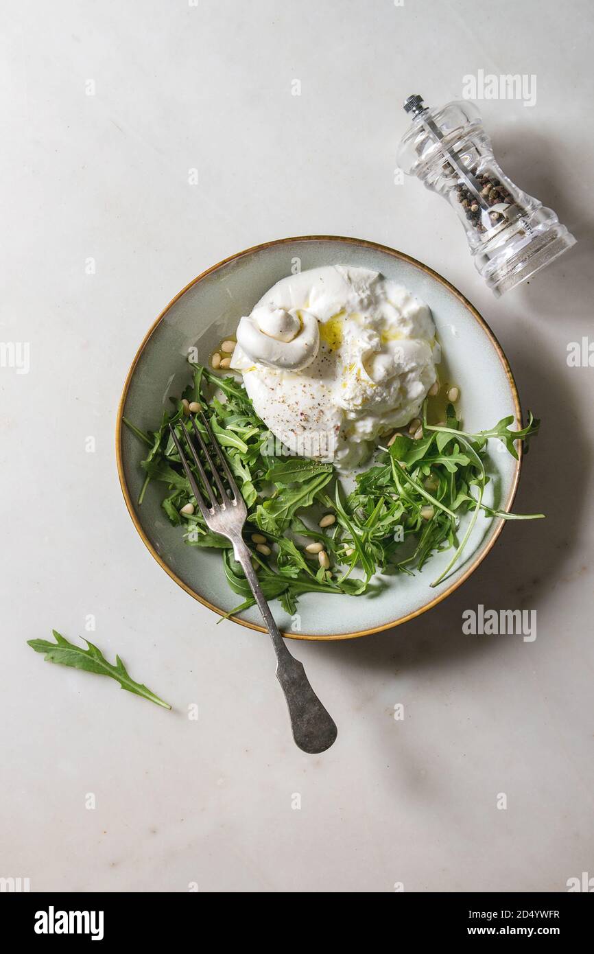 Geschnetzeltes Italienische burrata Käse, schwarzer Pfeffer Mühle, frische Rucola Salat, Pinienkerne und Olivenöl in weißer Keramik Platte über weißem Marmor Hintergrund. Fl Stockfoto