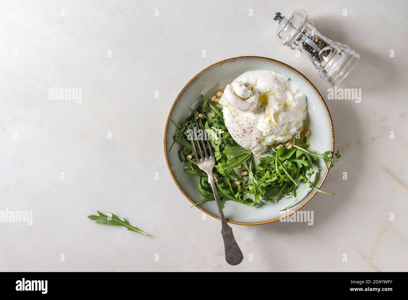 Geschnetzeltes Italienische burrata Käse, schwarzer Pfeffer Mühle, frische Rucola Salat, Pinienkerne und Olivenöl in weißer Keramik Platte über weißem Marmor Hintergrund. Fl Stockfoto