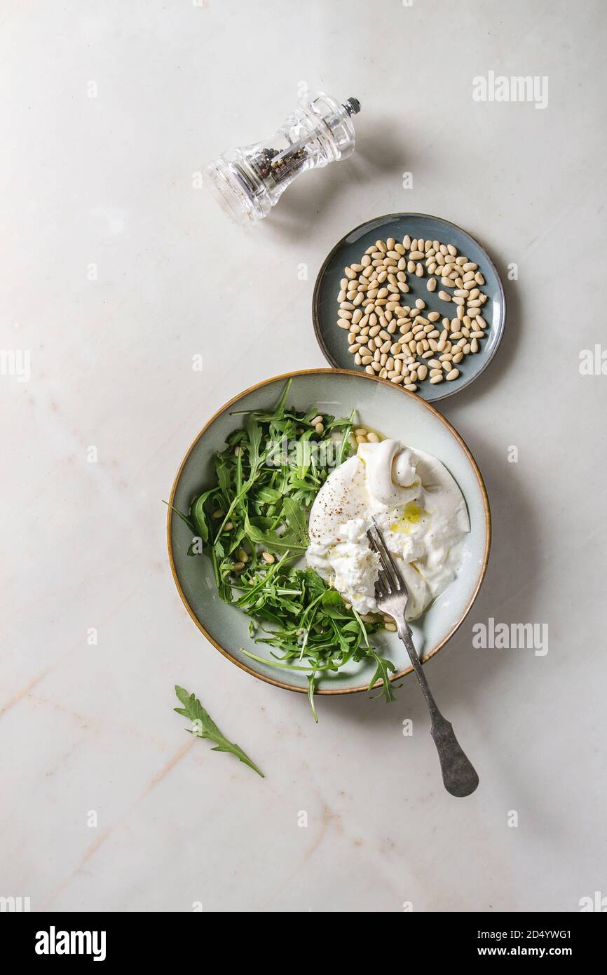 Geschnetzeltes Italienische burrata Käse, schwarzer Pfeffer Mühle, frische Rucola Salat, Pinienkerne und Olivenöl in weißer Keramik Platte über weißem Marmor Hintergrund. Fl Stockfoto