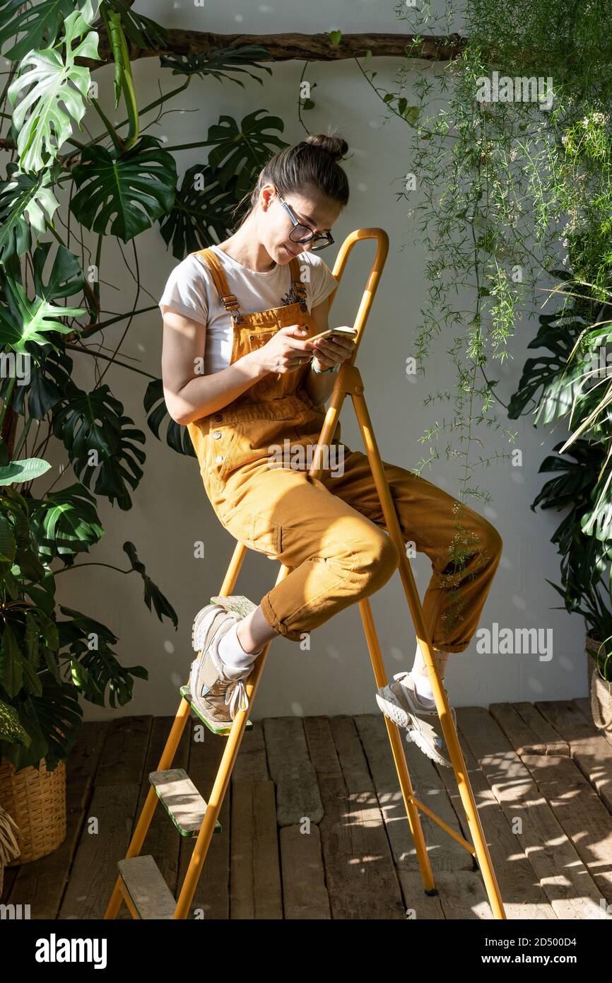 Gärtnerin in orangefarbenen Overalls sitzt auf der Trittleiter in ihrem grünen Haus, ruht sich aus, benutzt Smartphone, liest Nachricht am Telefon. Pause während der Arbeit Stockfoto