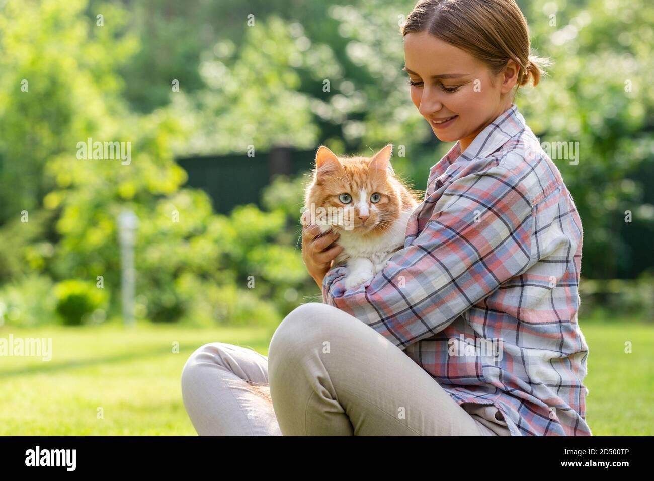 Nahaufnahme der lächelnden Frau in kariertem Hemd umarmt und umarmt mit Zärtlichkeit und Liebe Hausginger Katze, streichelt auf dem Kopf, im Freien in der Sonne Stockfoto