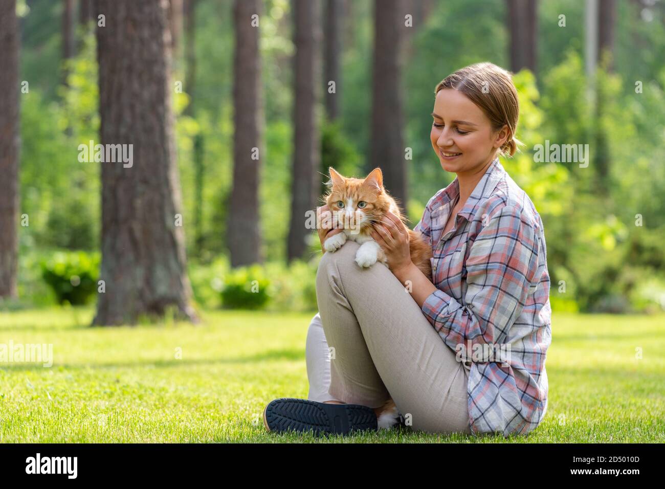 Nahaufnahme der lächelnden Frau in kariertem Hemd umarmt und umarmt mit Zärtlichkeit und Liebe Hausginger Katze, streichelt auf dem Kopf, im Freien in der Sonne Stockfoto