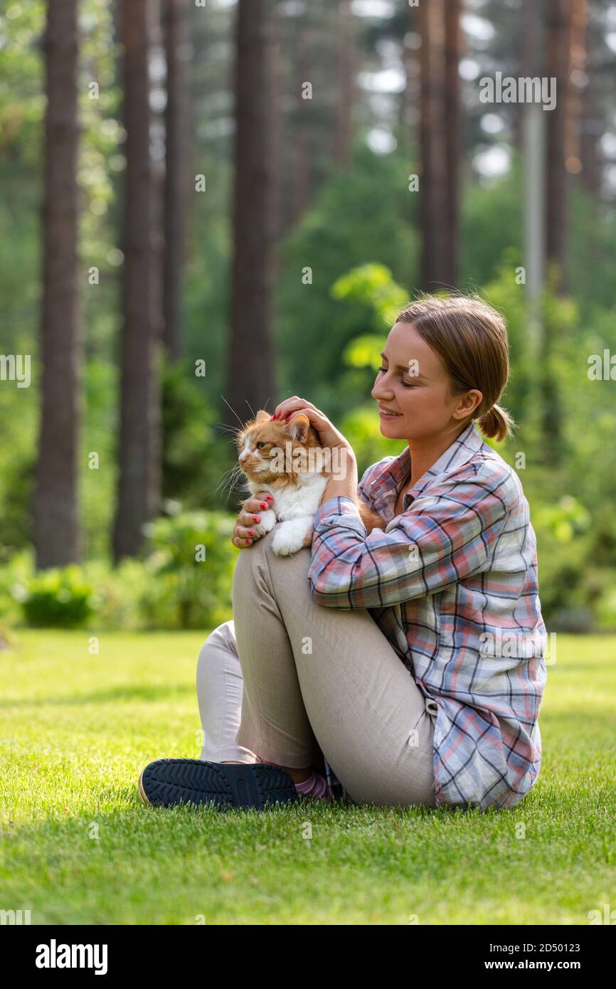 Glückliche Frau im karierten Hemd umarmt und umarmt mit Zärtlichkeit und Liebe Hausginger Katze, sitzt auf Gras im Freien an sonnigen Tag. Liebe zum A Stockfoto