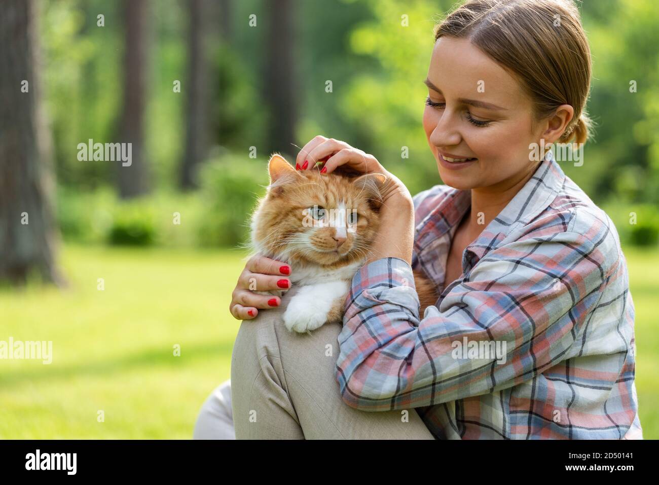 Nahaufnahme der lächelnden Frau in kariertem Hemd umarmt und umarmt mit Zärtlichkeit und Liebe Hausginger Katze, streichelt auf dem Kopf, im Freien in der Sonne Stockfoto