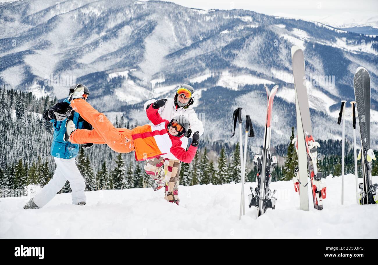Männliche Skifahrer in Skianzügen halten charmante Dame und werfen sie hoch in die Luft, Spaß auf verschneiten Hügel im Skigebiet mit schönen Bergen im Hintergrund. Konzept von Skifahren und Freundschaft. Stockfoto