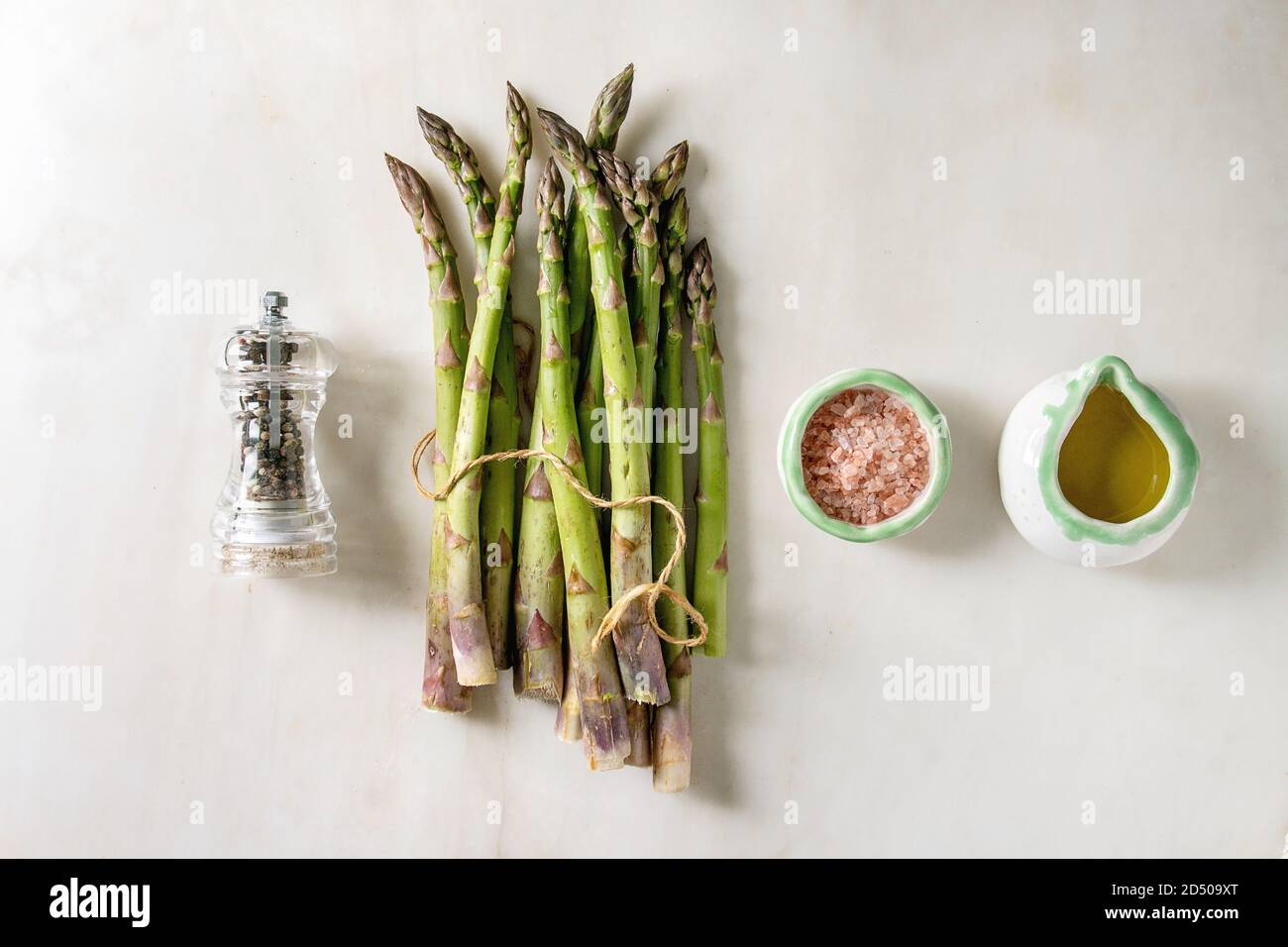 Bündel von organischen grüner Spargel mit Zutaten zum Kochen rosa Meersalz, schwarzer Pfeffer und Olivenöl in weißem Marmor Hintergrund. Flach, Raum Stockfoto