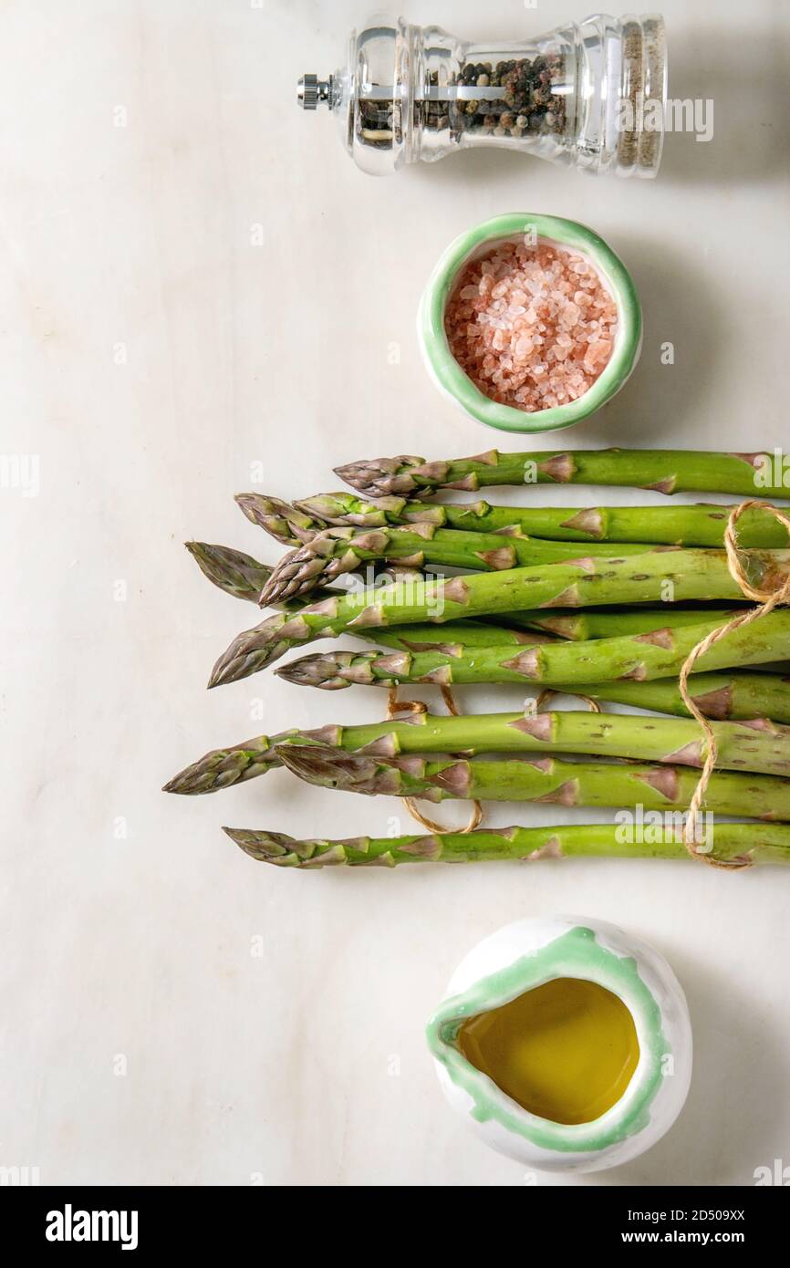 Bündel von organischen grüner Spargel mit Zutaten zum Kochen rosa Meersalz, schwarzer Pfeffer und Olivenöl in weißem Marmor Hintergrund. Flach, Raum Stockfoto