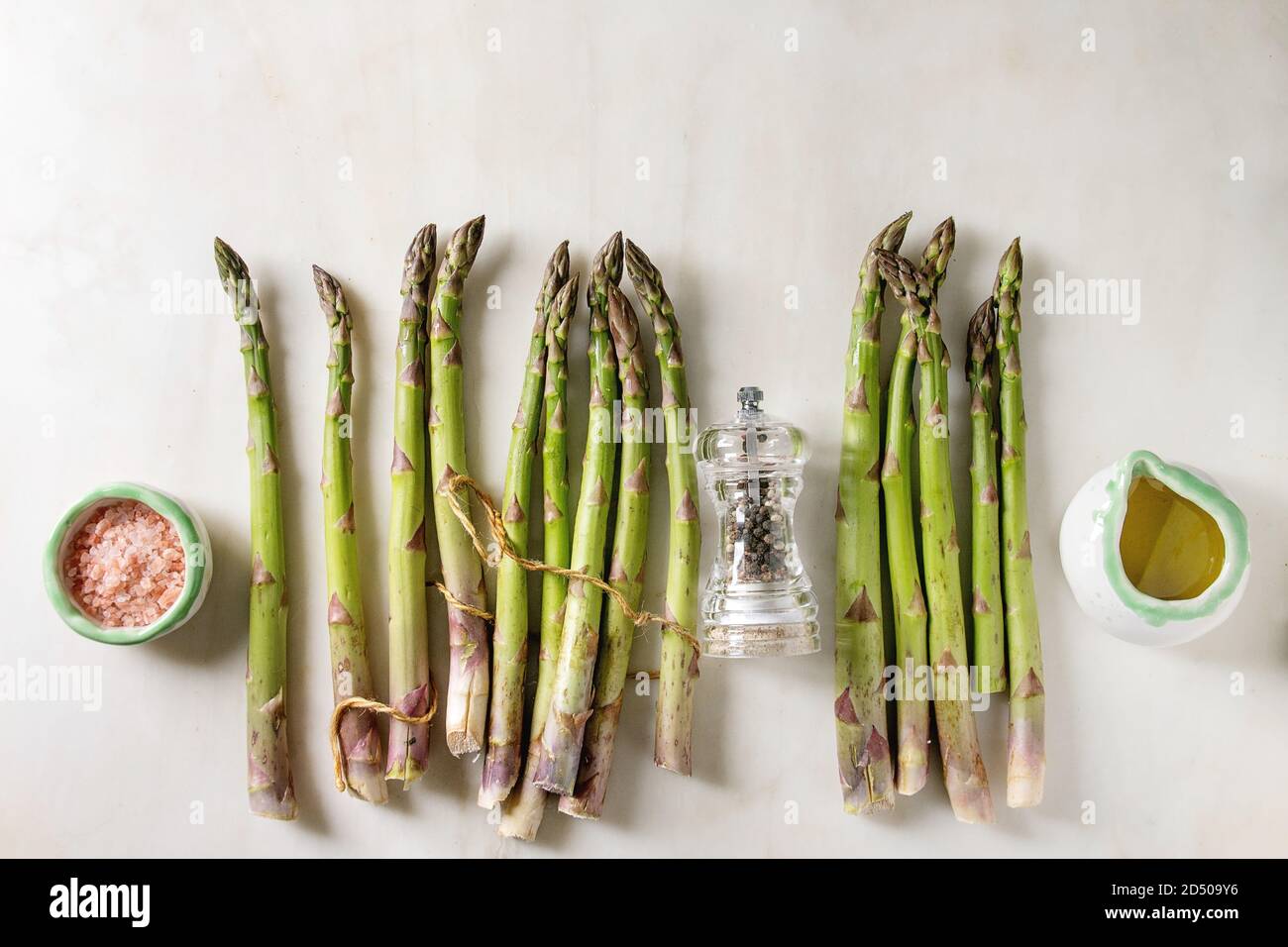 Bündel von organischen grüner Spargel mit Zutaten zum Kochen rosa Meersalz, schwarzer Pfeffer und Olivenöl in weißem Marmor Hintergrund. Flach, Raum Stockfoto