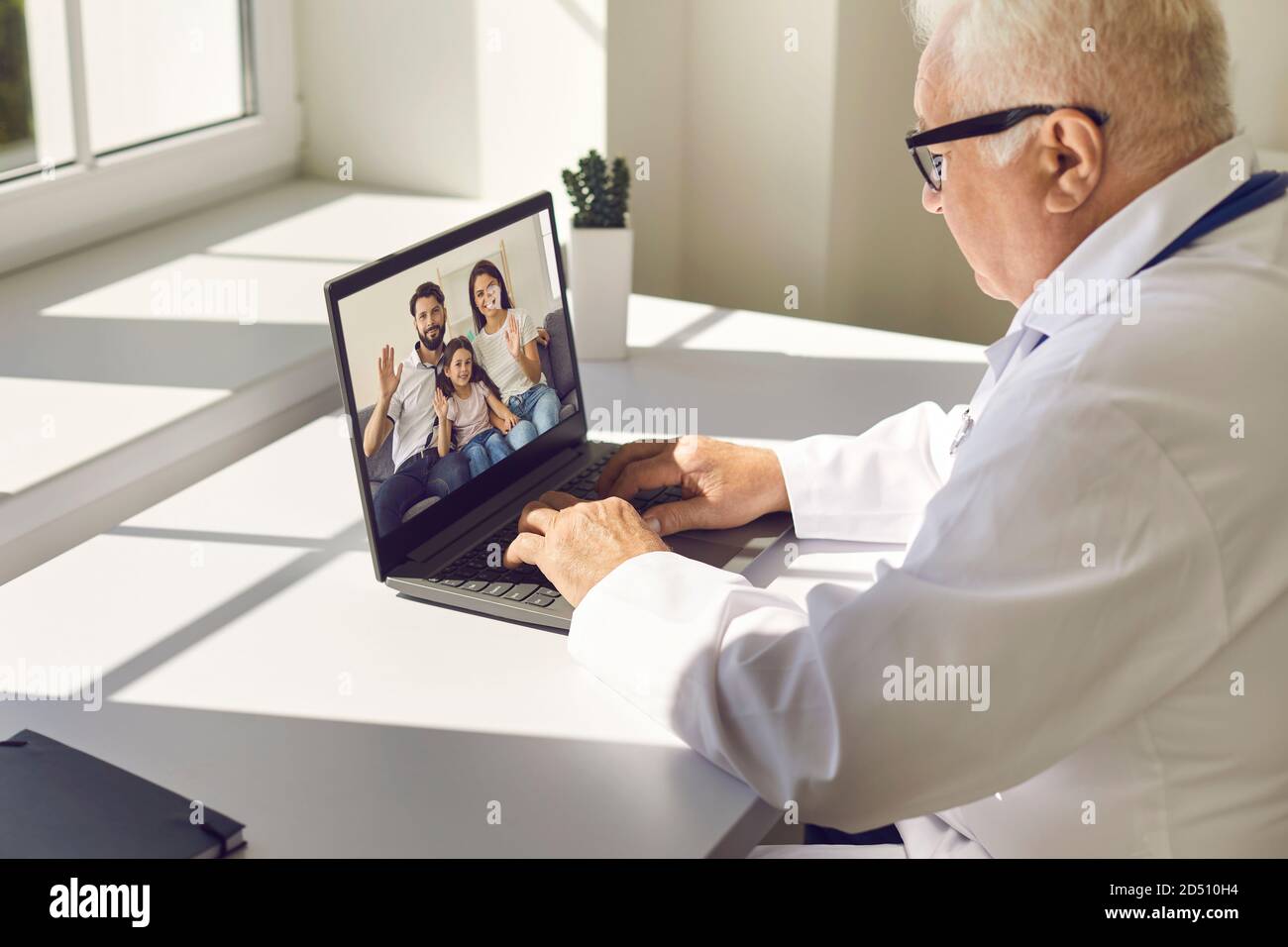 Reifer Oberarzt in Uniform sitzt am Laptop und kommuniziert Mit jungen lächelnden Familie Stockfoto