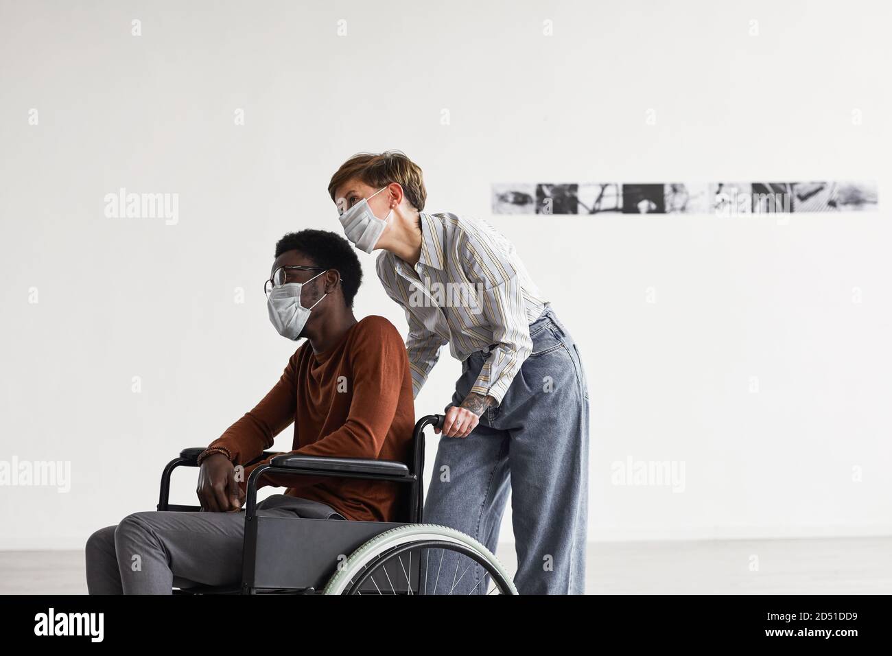 Minimalporträt eines afroamerikanischen Mannes, der Rollstuhl benutzt und Gemälde in der Galerie moderner Kunst ansieht, wobei ihm eine junge Frau hilft, die beide Masken trägt, Raum kopieren Stockfoto