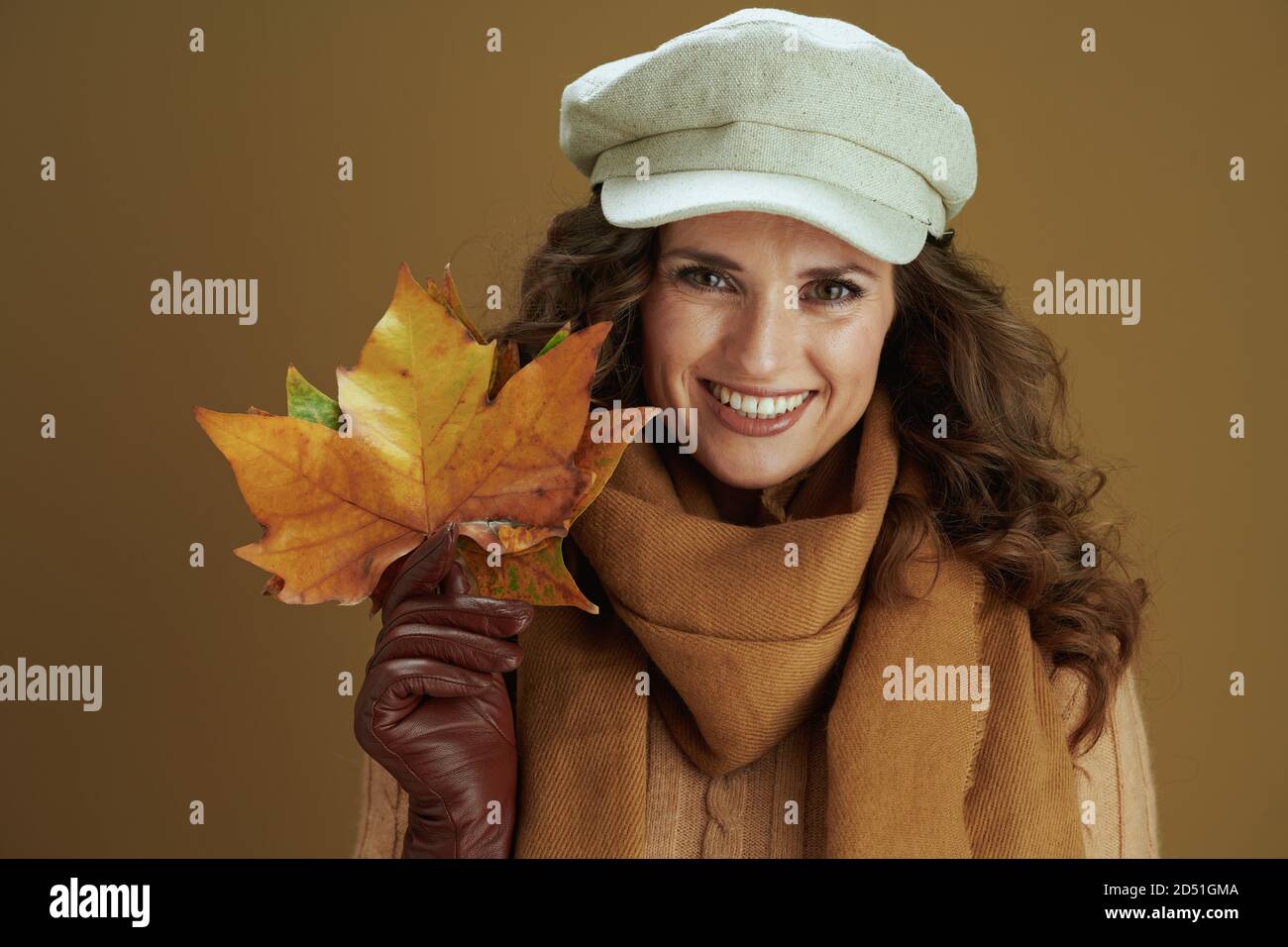 Hallo Herbst. Portrait der lächelnden modernen 40 Jahre alten Frau in Pullover mit Lederhandschuhen und gelben Herbst Ahornblätter isoliert auf beige. Stockfoto