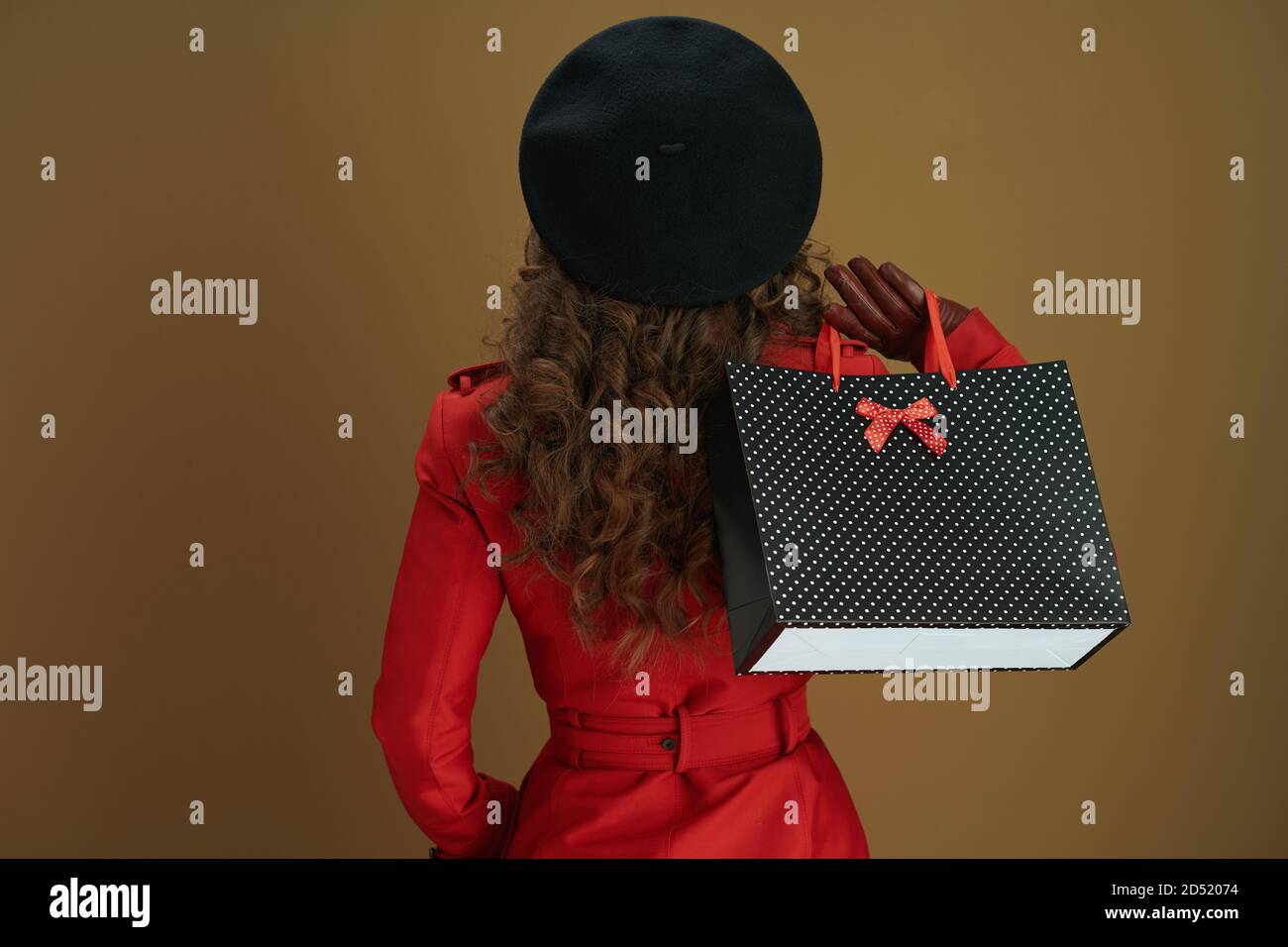 Hallo november. Von hinten gesehen Frau in rotem Mantel und schwarzer Baskenmütze mit Lederhandschuhen und Papier Einkaufstasche isoliert auf Bronze Hintergrund. Stockfoto