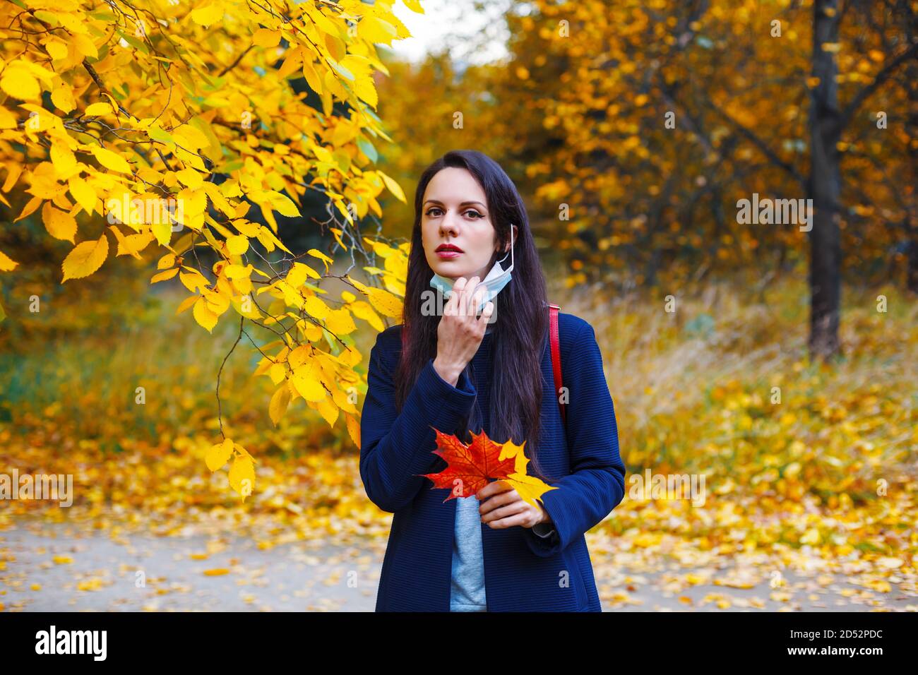 Brunette kaukasische Frau zieht nach unten Medizin Maske im bunten Herbst Park. Stockfoto