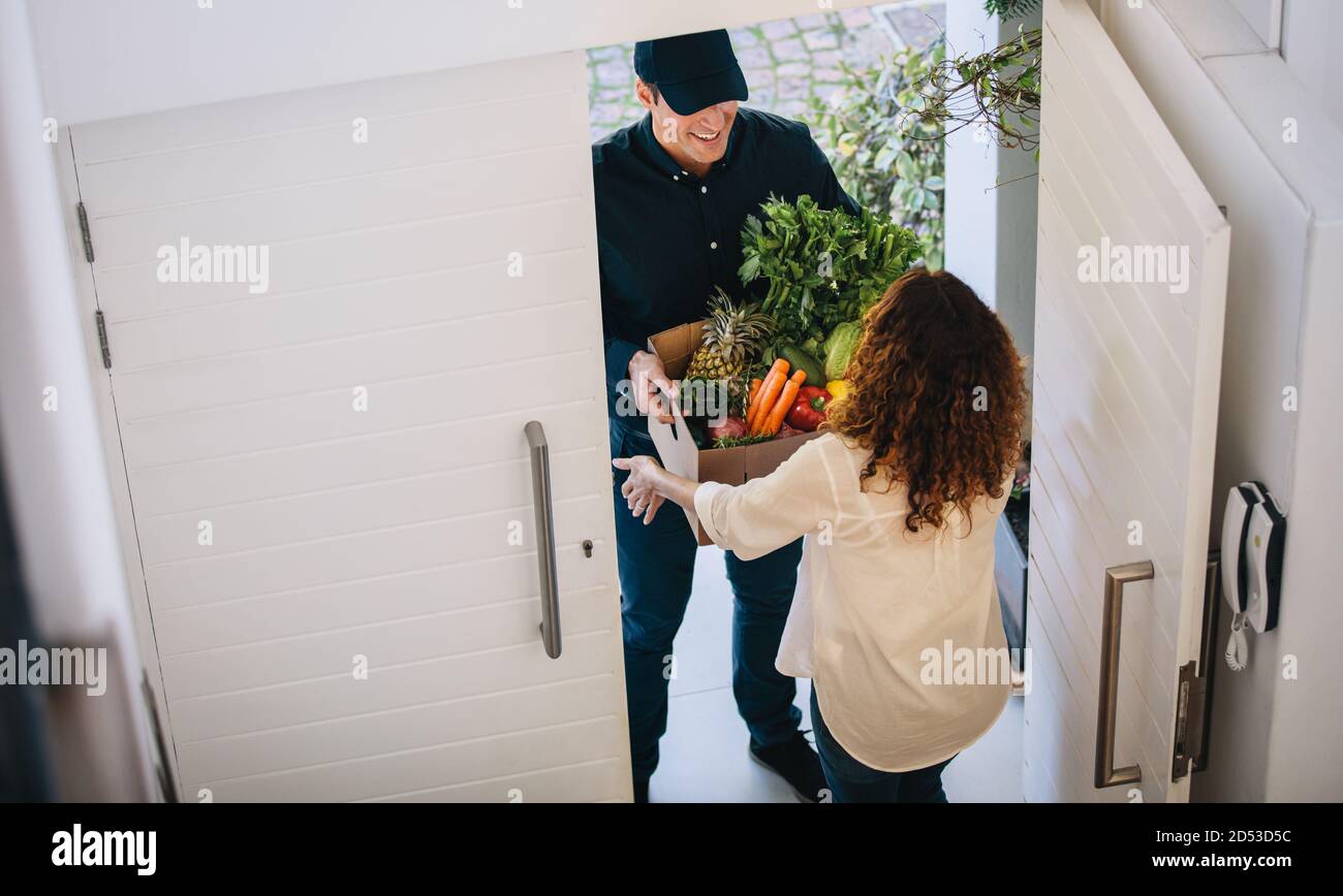 Blick aus der Perspektive einer Kundin, die Lebensmittel vom Lieferer vor der Haustüre erhält. Liefermitarbeiter liefert eine Schachtel Obst und Gemüse Stockfoto