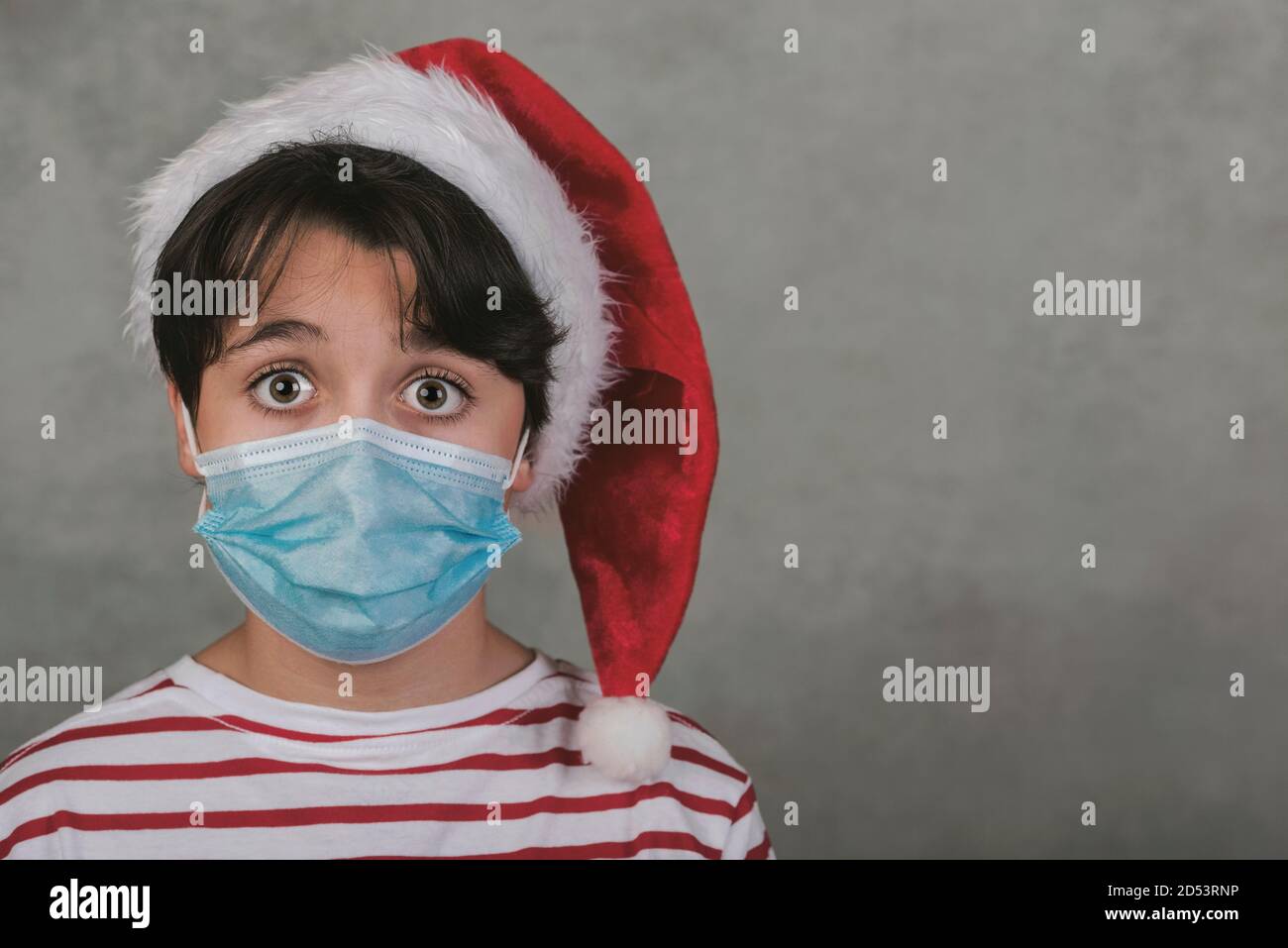 Frohe Weihnachten, Nahaufnahme des Kindes mit medizinischer Maske trägt Weihnachtsmann hut auf grauem Hintergrund Stockfoto
