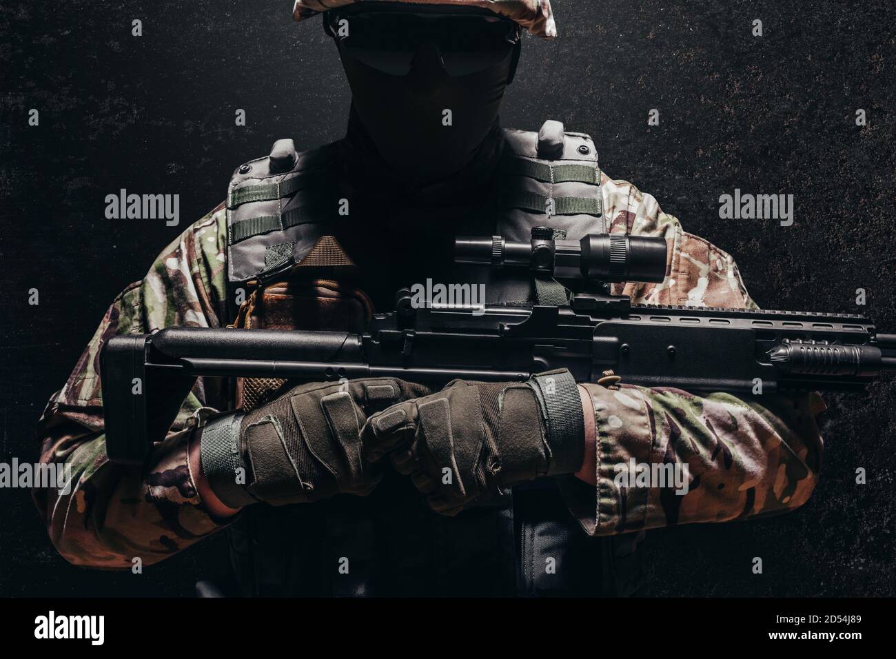 Foto eines voll ausgestatteten schattigen Soldaten im Helm, Panzerweste mit Scharfschützengewehr auf dunklem Hintergrund. Stockfoto
