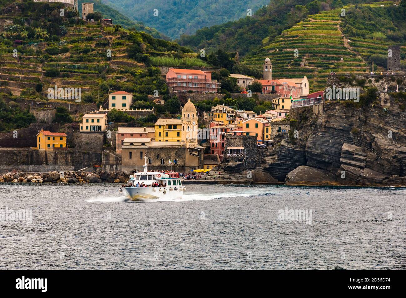 Tolle Nahaufnahme von Vernazza vom Meer aus. Ein Schiff mit Besuchern verlässt den Hafen. Im Hintergrund sind bunte Häuser, die Kirche von Santa... Stockfoto