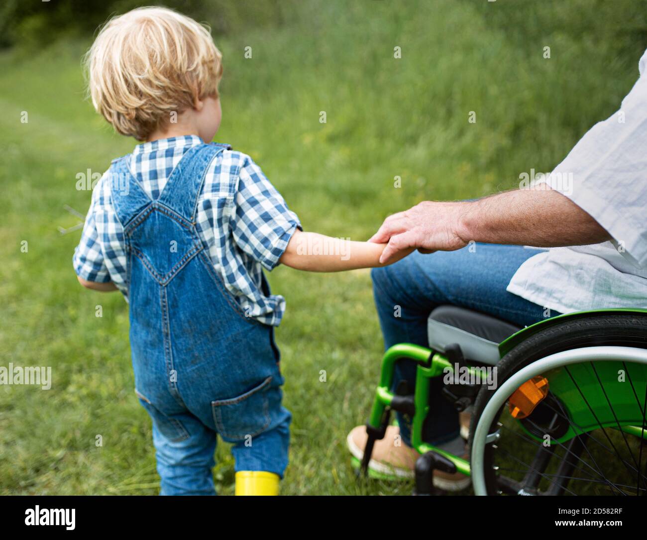 Kleiner Junge mit nicht erkennbarem Senioren Großvater im Rollstuhl auf einem Spaziergang auf der Wiese in der Natur. Stockfoto