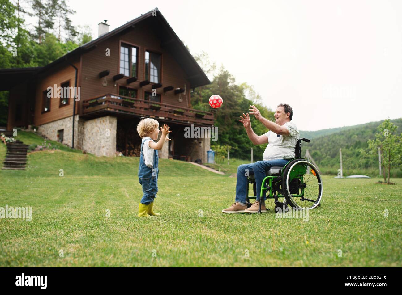 Kleiner Junge mit Seniorengroßvater im Rollstuhl im Garten, spielt mit einem Ball. Stockfoto